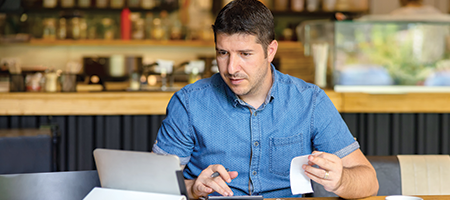 male business owner sitting at counter with laptop keying in receipts