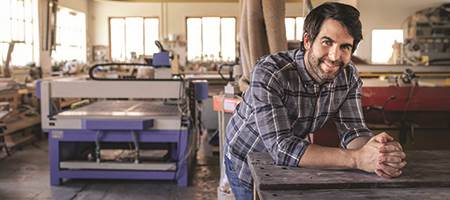 smiling male business owner leaning on workbench in manfacturing shop